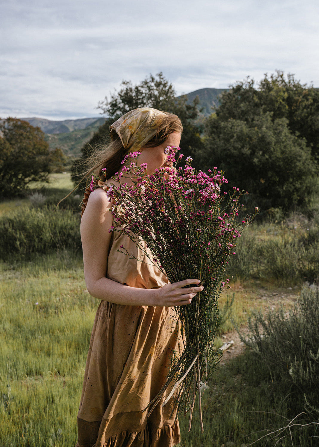 Woman holding a bundle of flowers in a natural setting with trees and mountains in the background