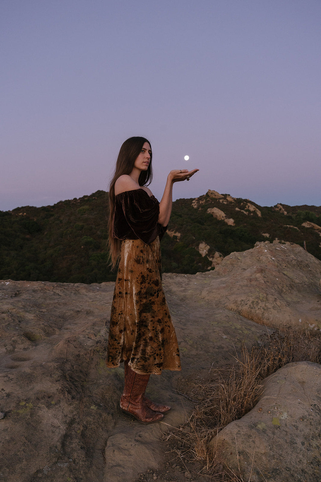Woman in a patterned dress holding the moon in her hand against a mountainous landscape.