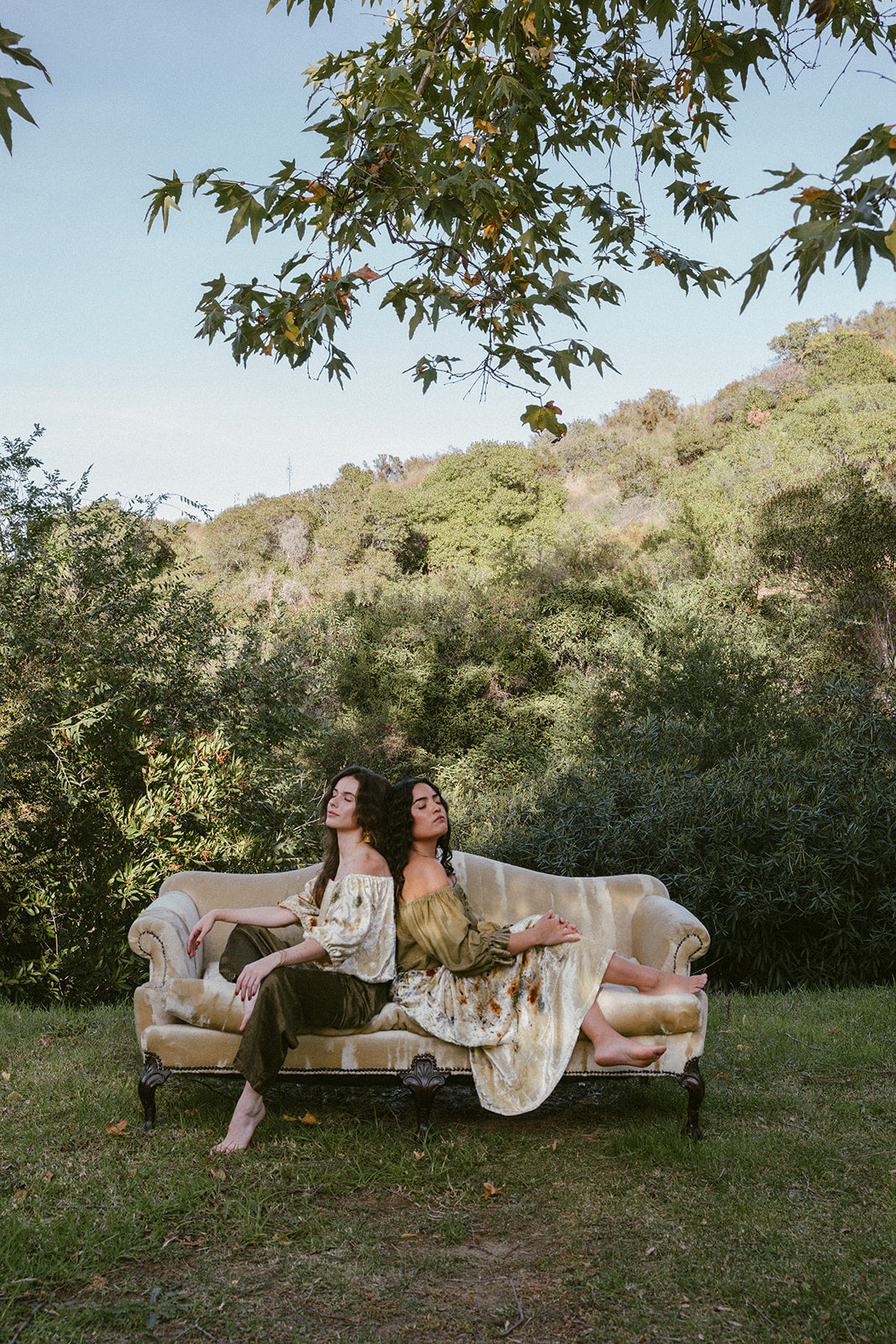 Two women sitting on a vintage sofa in a forest setting
