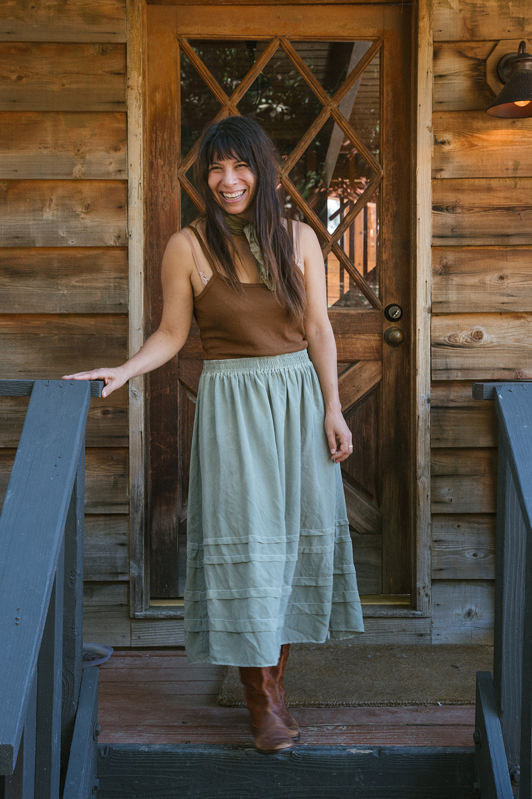 Woman standing on a wooden porch wearing a brown top and light green skirt.