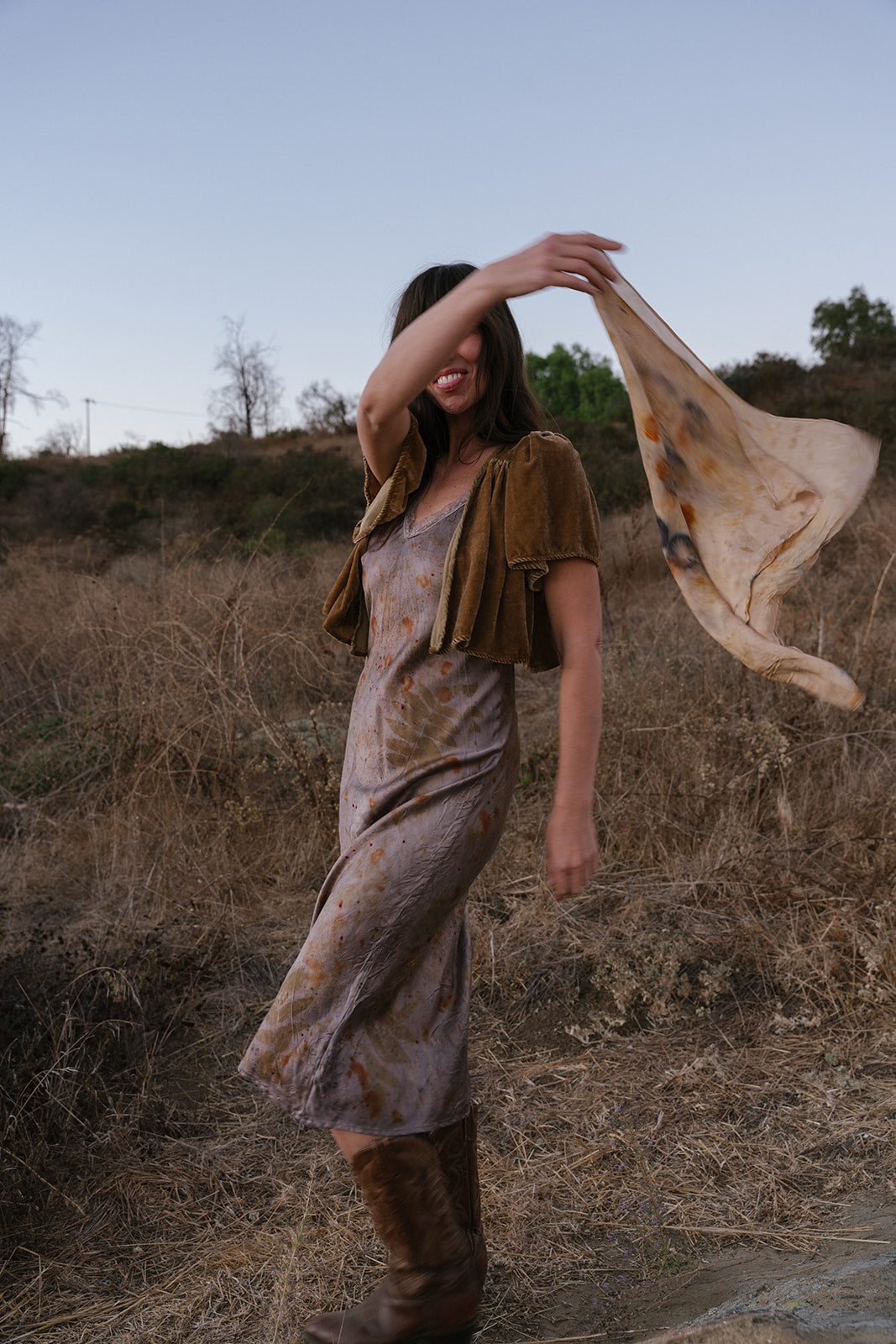 Woman in a field holding a piece of fabric, wearing a dress and boots.