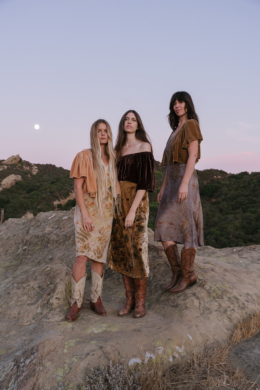 Three women standing on a rocky outcrop with a mountainous landscape and sunset sky.