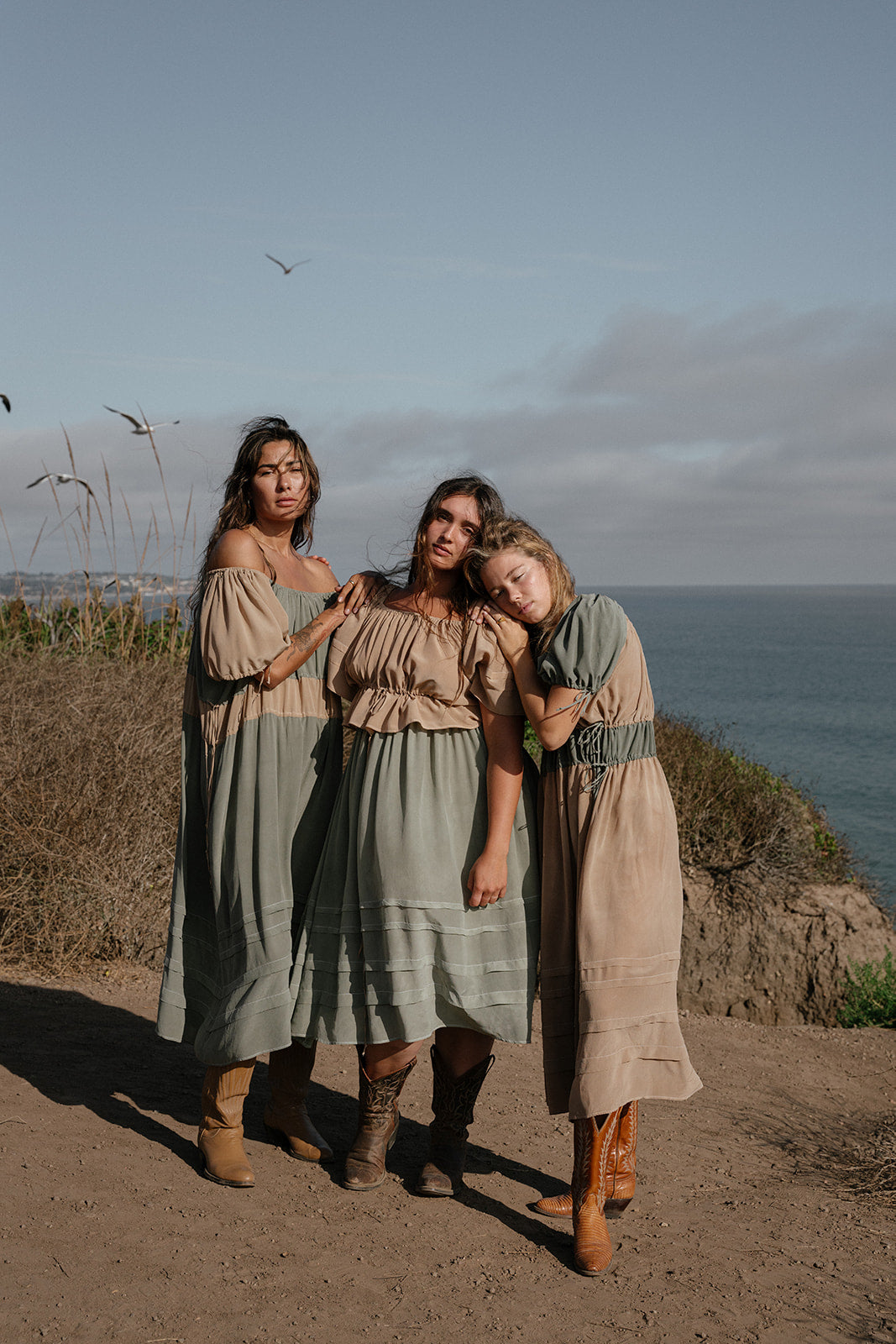 Three women in vintage-style dresses standing on a coastal cliff with ocean view.