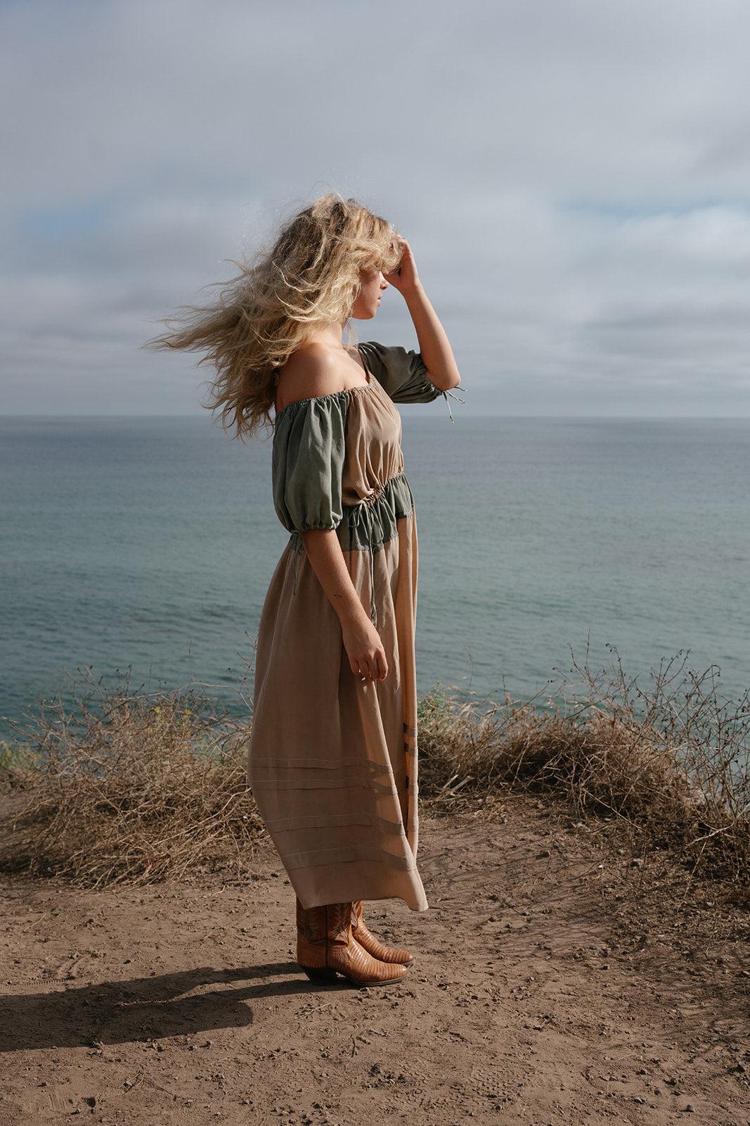 Woman in a long dress standing on a coastal path with ocean and sky in the background